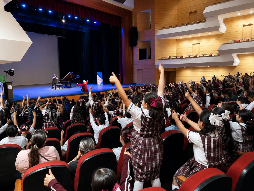 Un auditorio lleno de escolares con los brazos en alto, respondiendo al intérprete en el escenario.